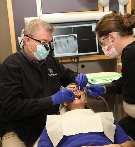 Dr. Larry Schmakel, in loupes and a mask, treating a patient with his assistant beside him.